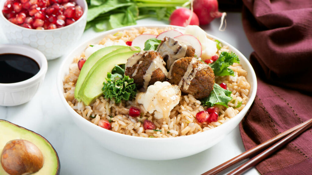 White bowl filled with fried rice, kale, deep fried falafel, avocado and pomegranate seeds topped with tahini sauce